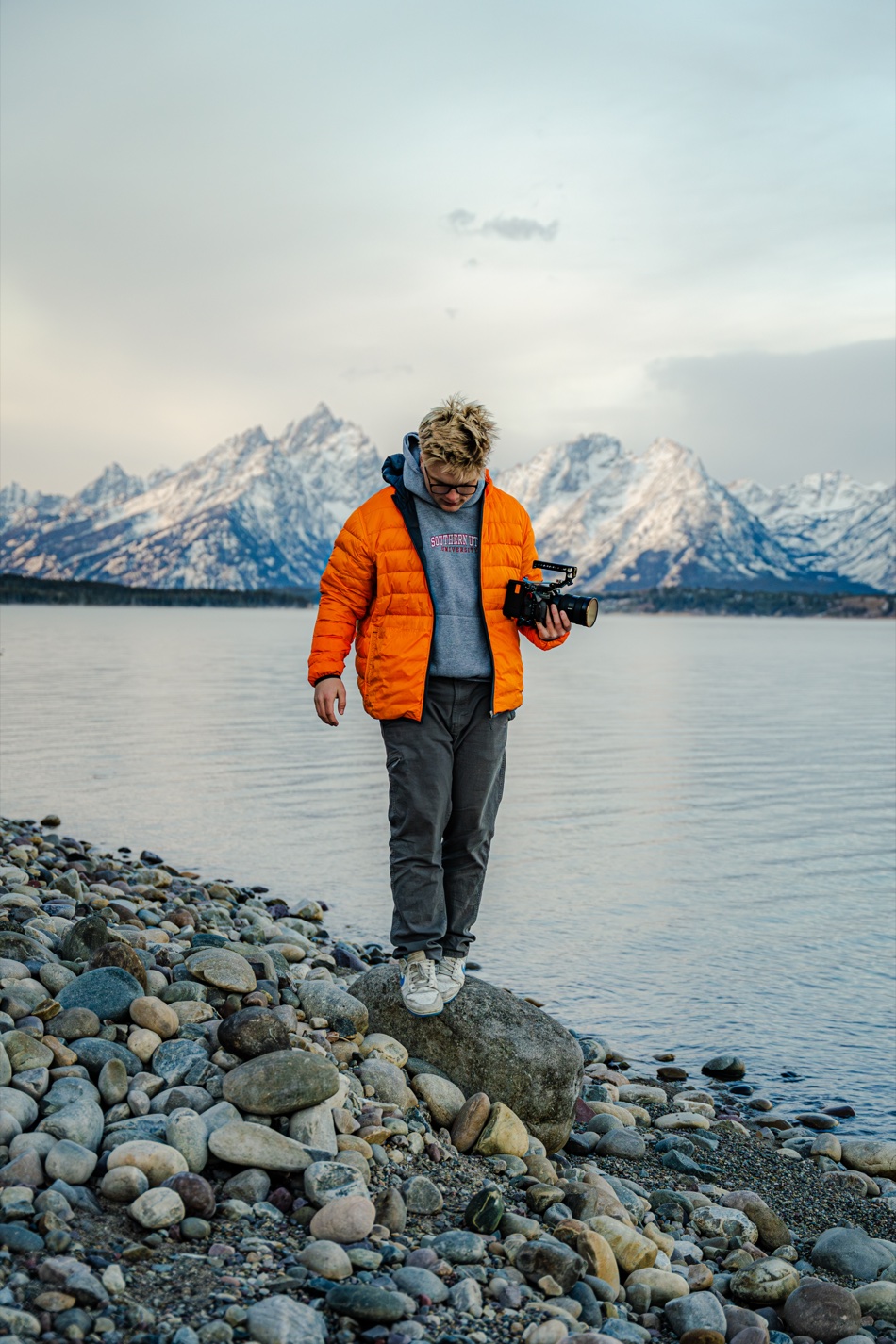 Ethan Dorny with a camera on the shore of Jackson Lake, Teton range behind